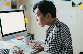 [Featured image] A young Asian man in a checkered shirt sits in front of a desktop computer and a laptop computer. 