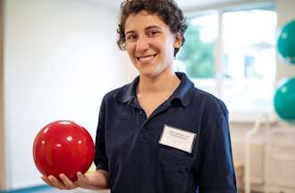 [Featured image] A physical therapist holding a red therapy ball stands in the middle of a clinic smiling at the camera.