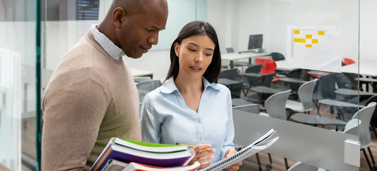 [FEATURED IMAGE] An MBA student holding a notebook asks a professor a question while standing outside a classroom.

