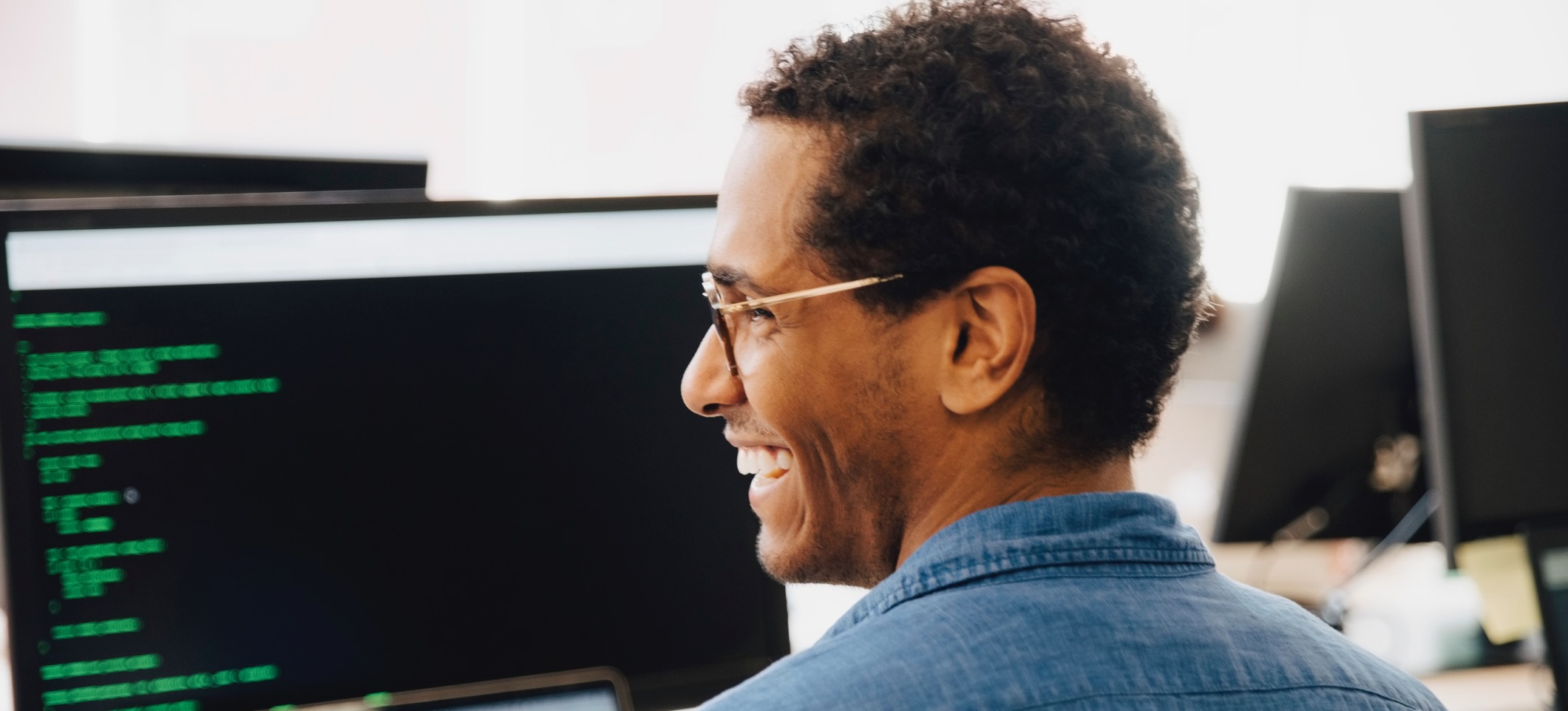 [Featured Image] A software engineer smiles in front of a computer monitor with code on the screen. 