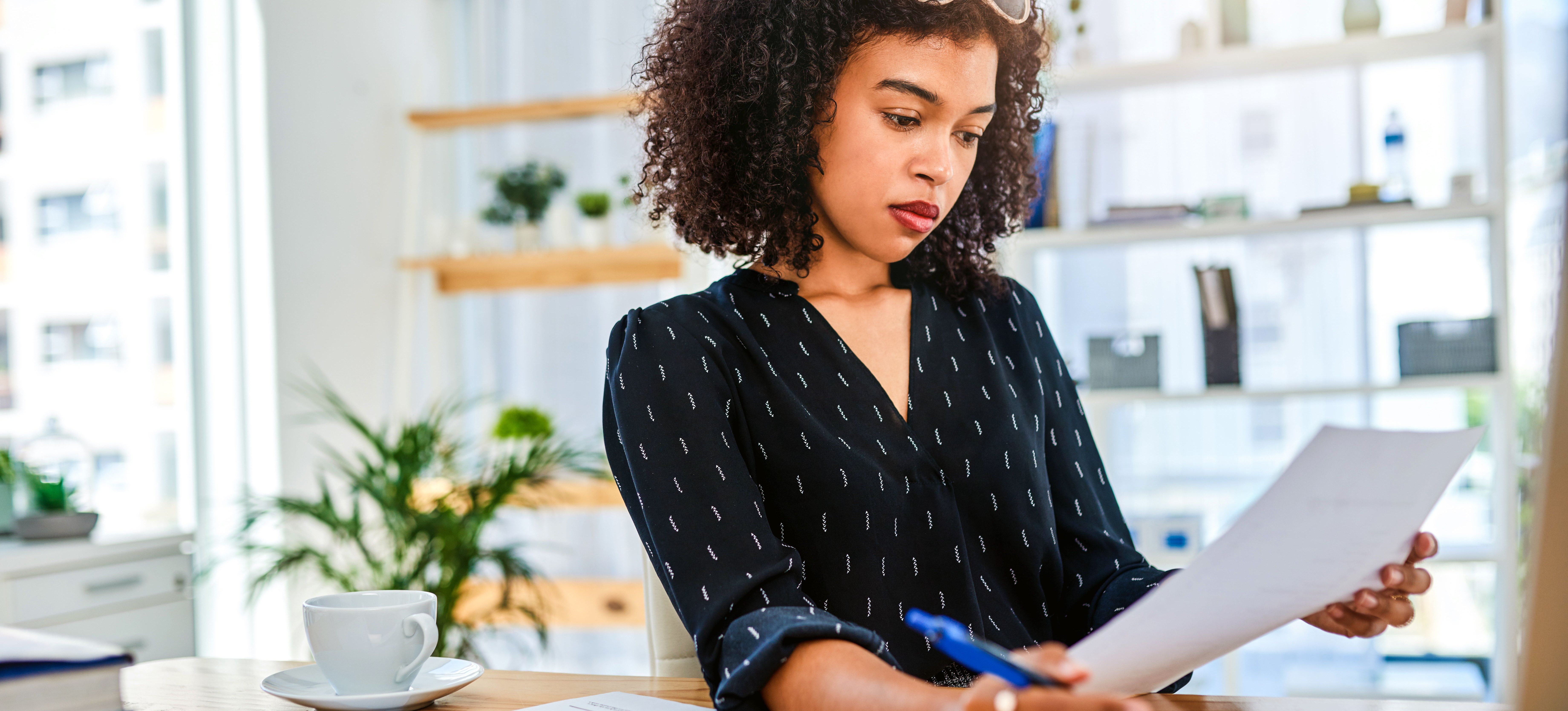 [Featured Image] A person is working at home, adding their GPA to their resume while standing at a desk with papers laid out, a cup of coffee, and a tablet.  