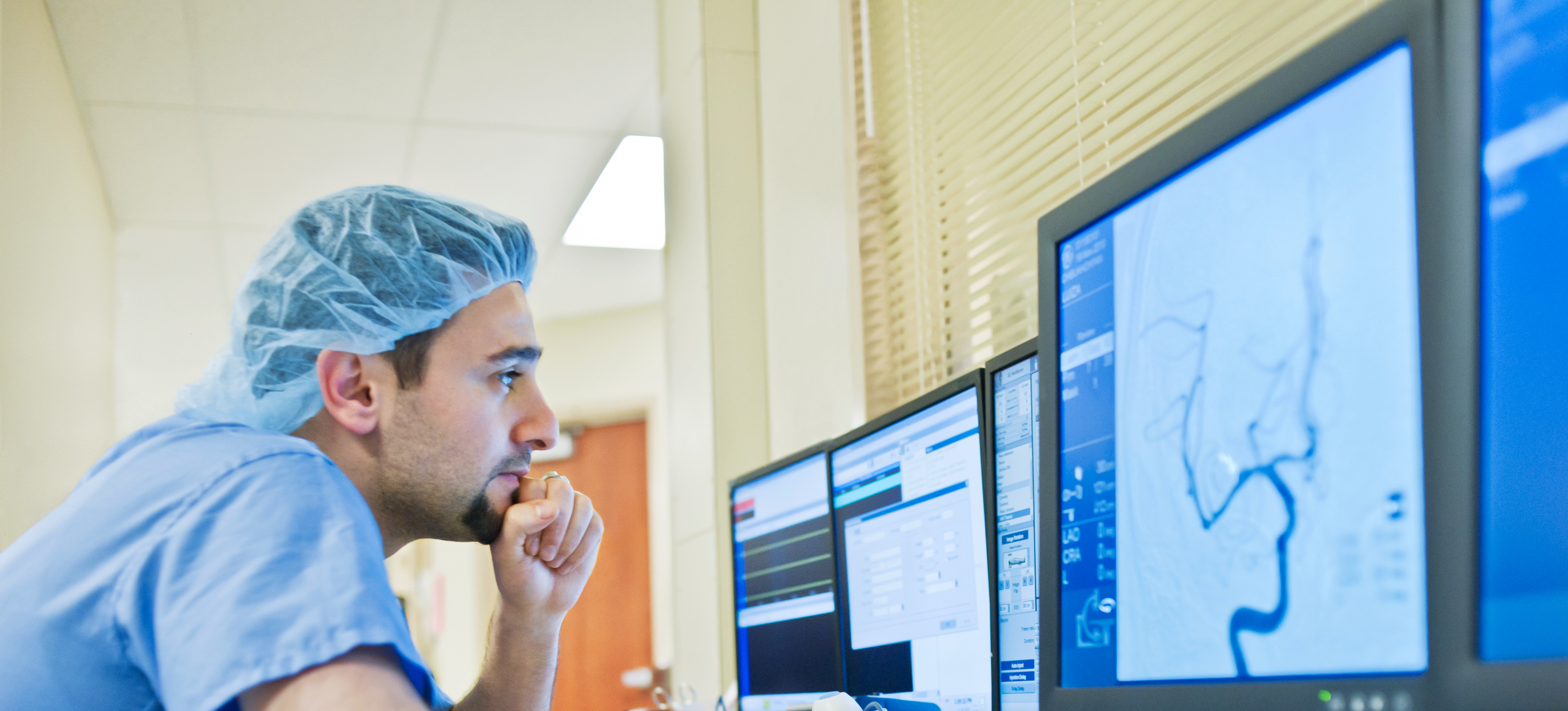 [Featured Image]: An epidemiologist in scrubs is studying and analyzing information on computer monitors to find the cause of a disease. 