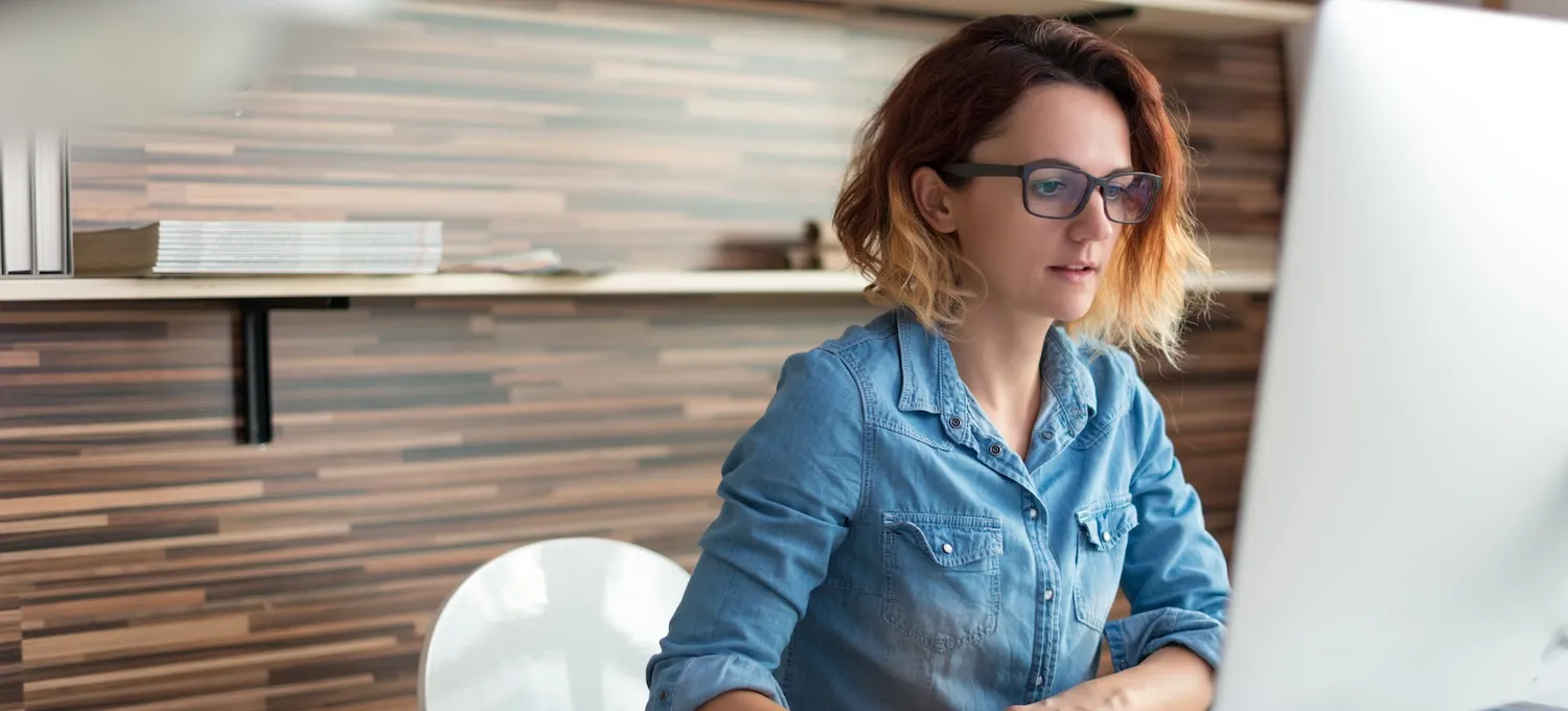 [Featured image] A person wearing glasses and sitting in an office uses their desktop to research whether they can earn their MBA without a bachelor's.
