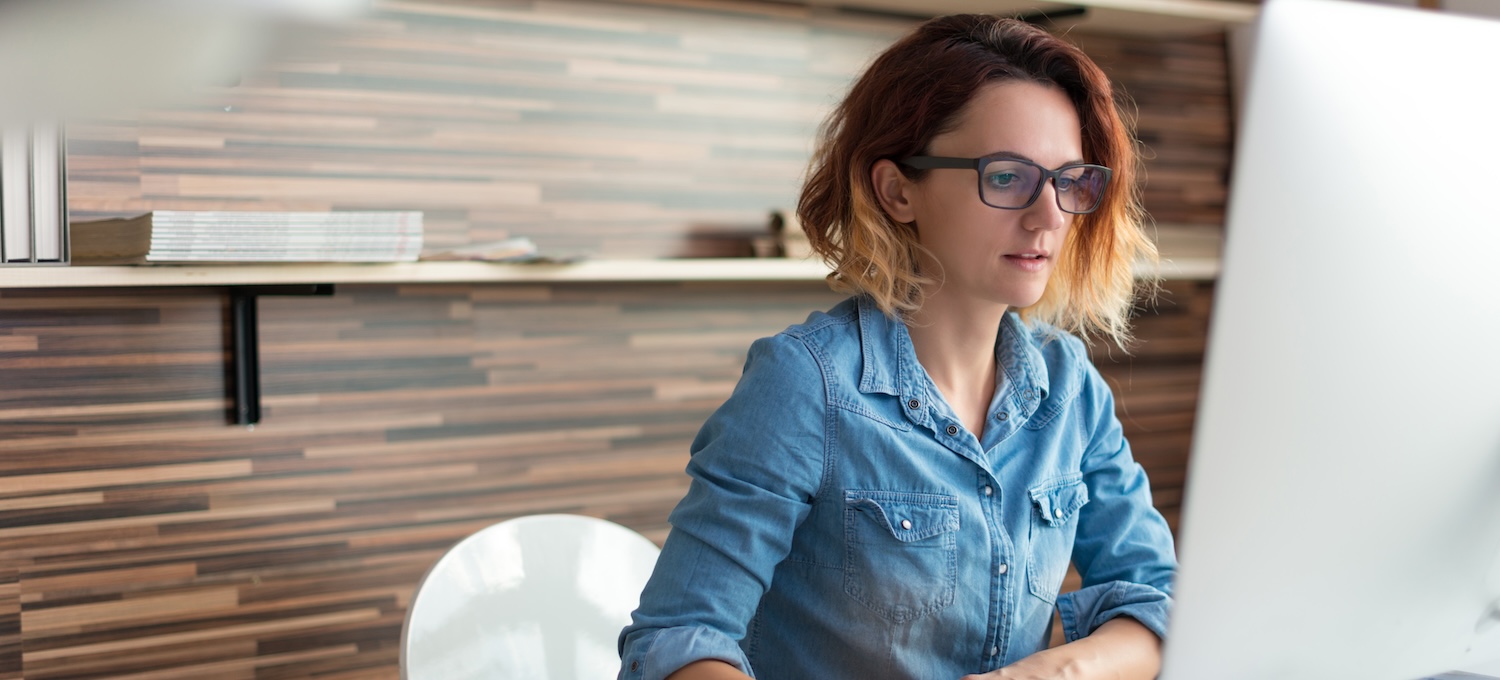[Featured image] A woman wearing glasses and sitting in an office uses her desktop to research whether she can earn her MBA without a bachelor's.