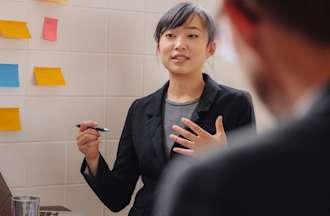 [Featured image] A young Asian woman dressed professionally stands speaking to two people shown in profile. 