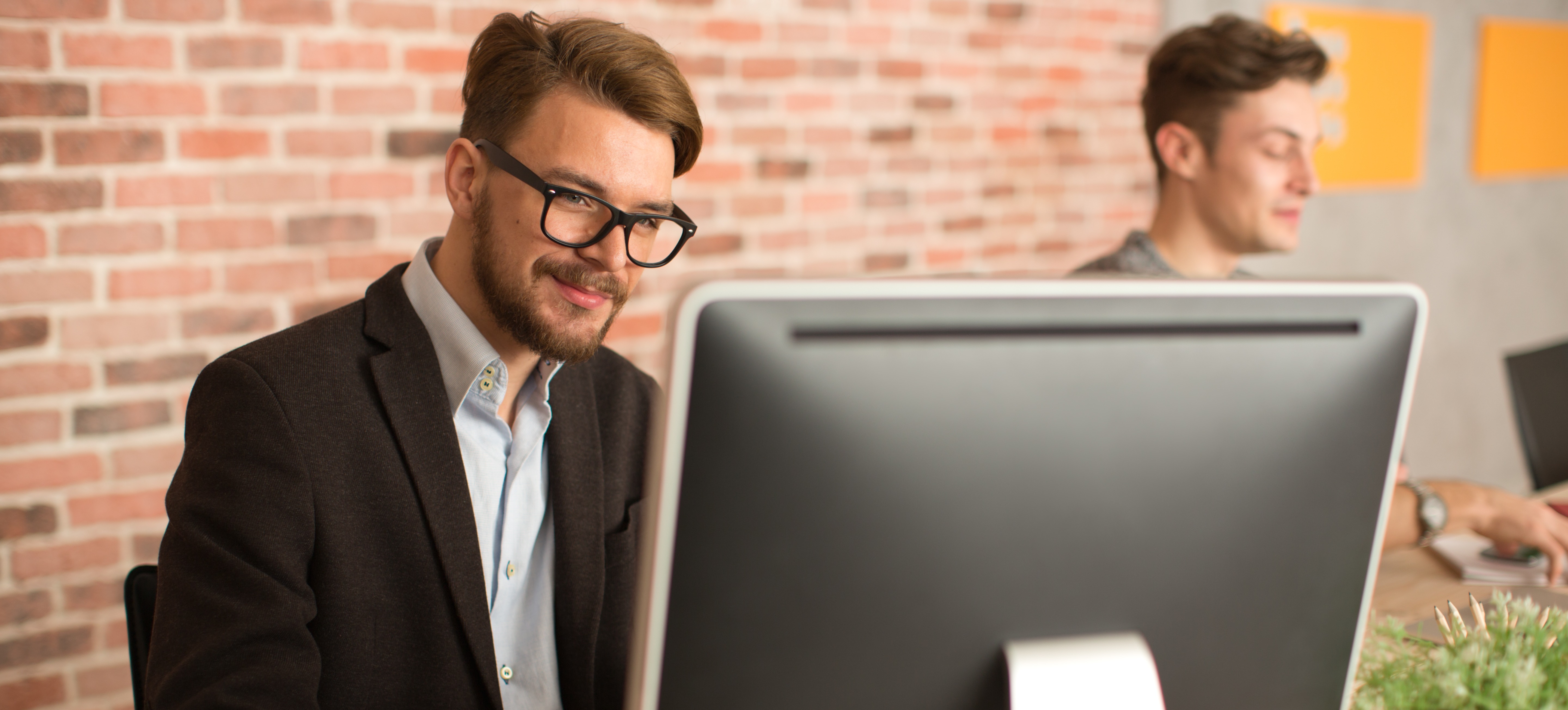[Feature Image] A marketing professional in a black jacket and dark-rimmed glasses performs Google Ads A/B testing on a desktop computer in a professional setting with a brick wall and a colleague in the background. 
