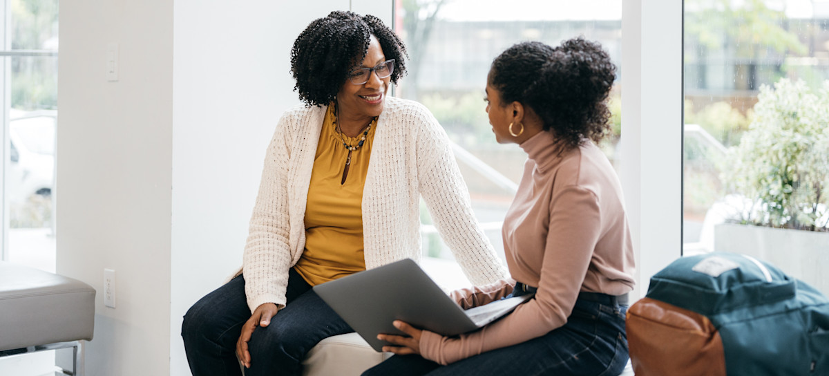 [Featured Image] A young student holding a laptop meets with a professor to discuss whether they should pursue a career in business intelligence vs. data analytics. 
