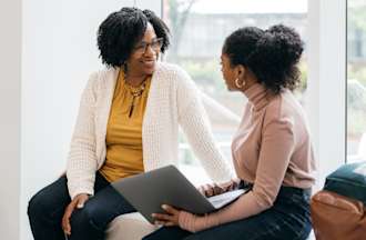 [Featured Image] A young student holding a laptop meets with a professor to discuss whether she should pursue a career in business intelligence vs. data analytics. 
