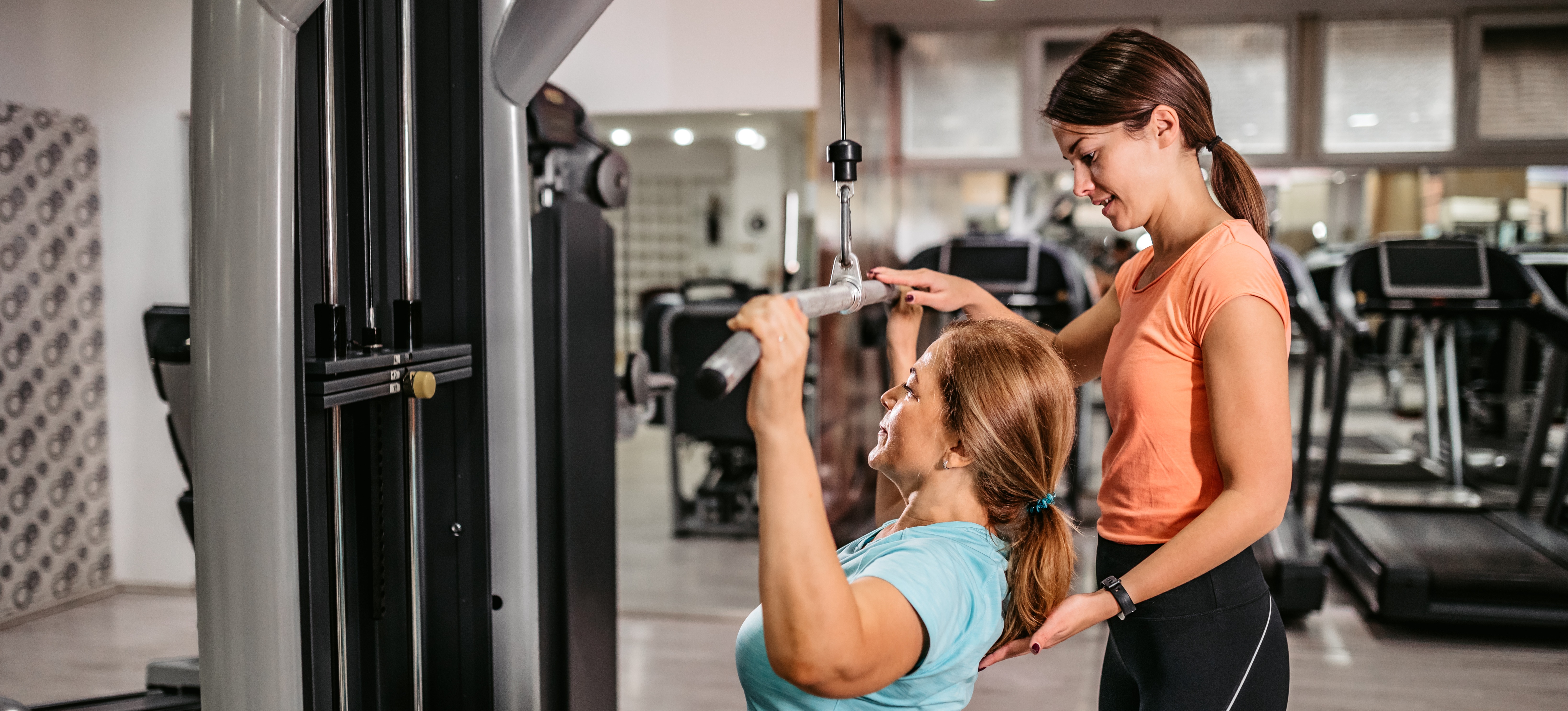 [Featured Image] A personal trainer helps an individual perfect their form on a weight machine.
