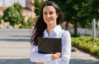 [Featured image] A young white woman with long brown hair, wearing a white collared shirt, stands on a college campus holding a folder. 