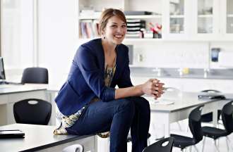 [Featured Image] A woman is sitting on a desk in a classroom wearing business casual clothing.