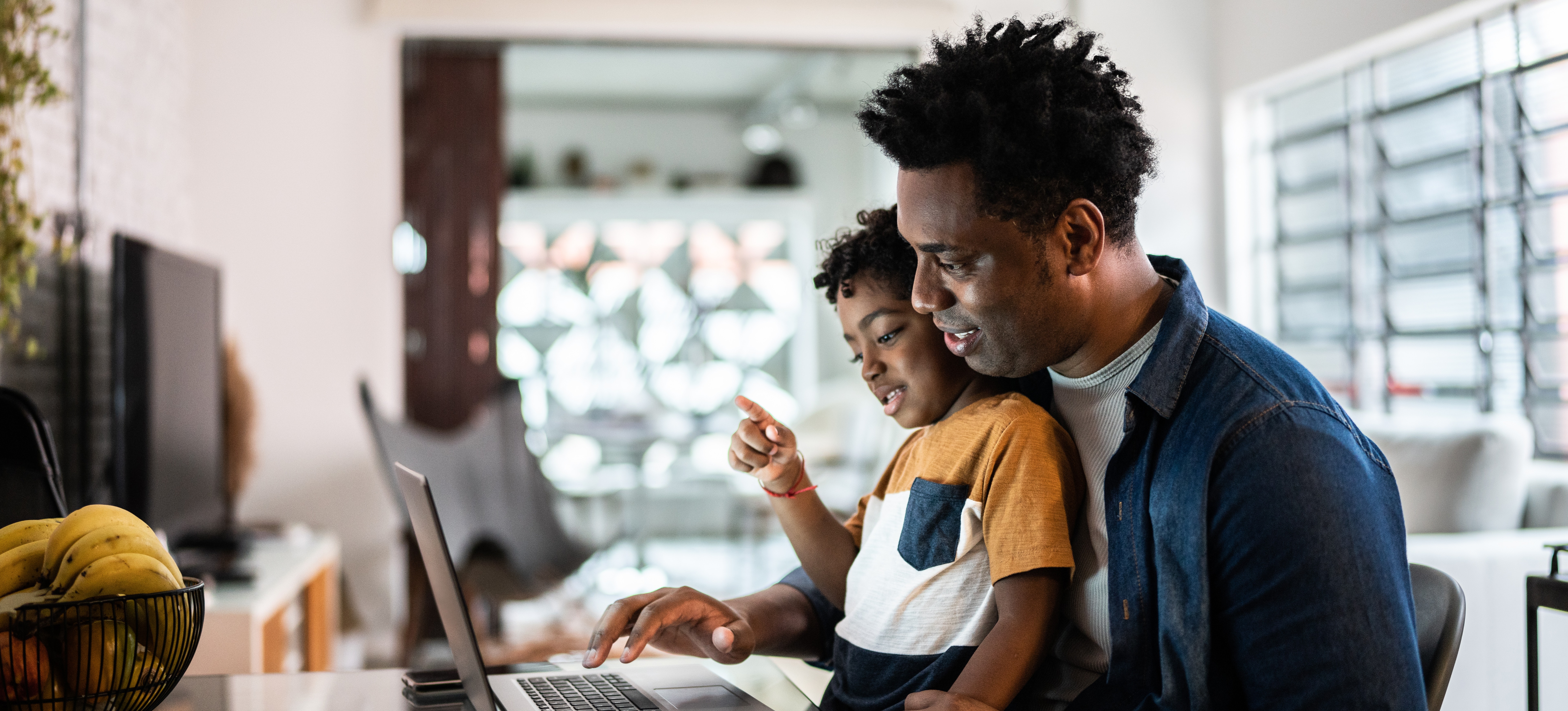[Featured image] A young parent sits at their laptop in their home and learns how to search in Google Sheets as their young child sits on their lap and watches the laptop screen.
