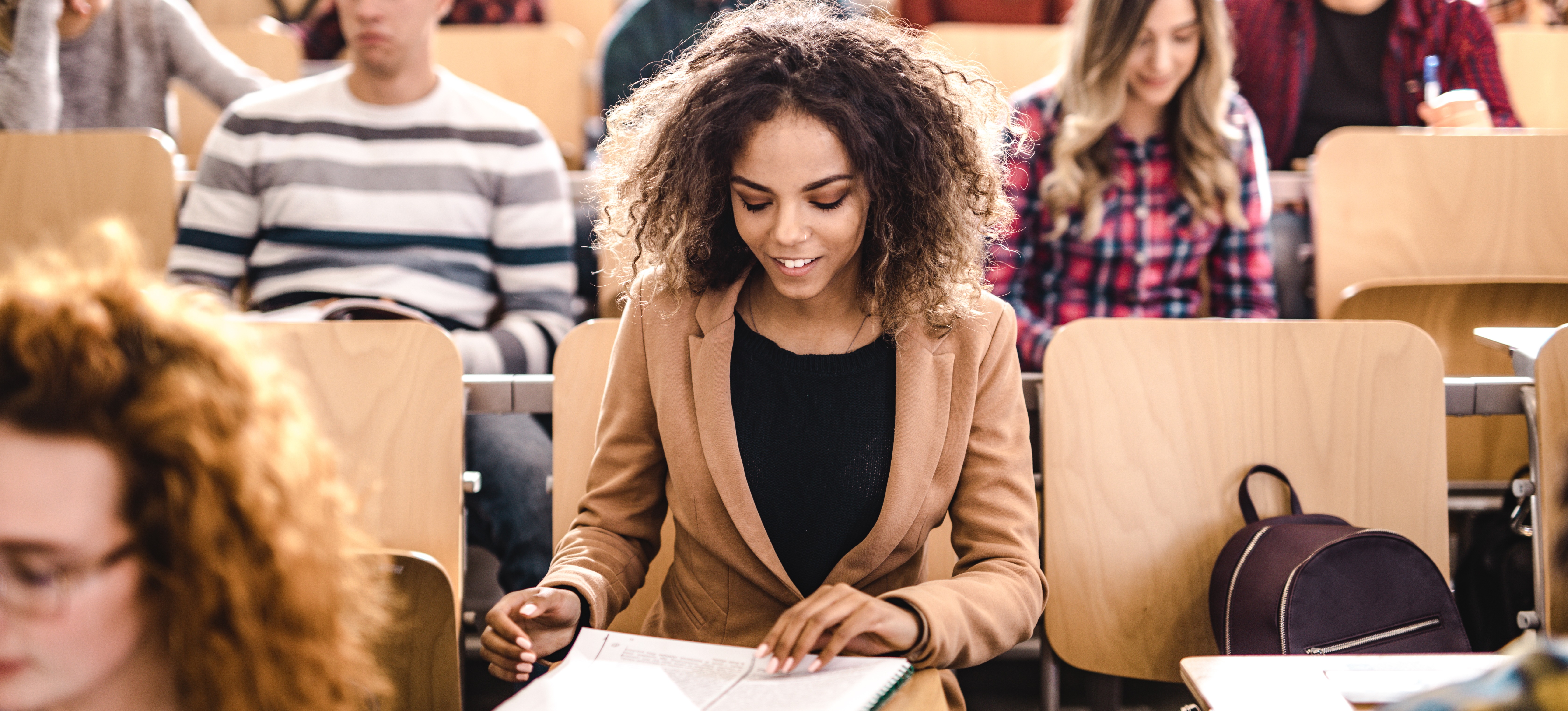 [Featured image] A young woman sits in a college classroom, looking at her notebook. 