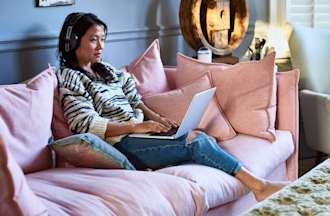 [Featured image] A website designer sits on a pink sofa in a living room with her computer on her lap working on a website design.