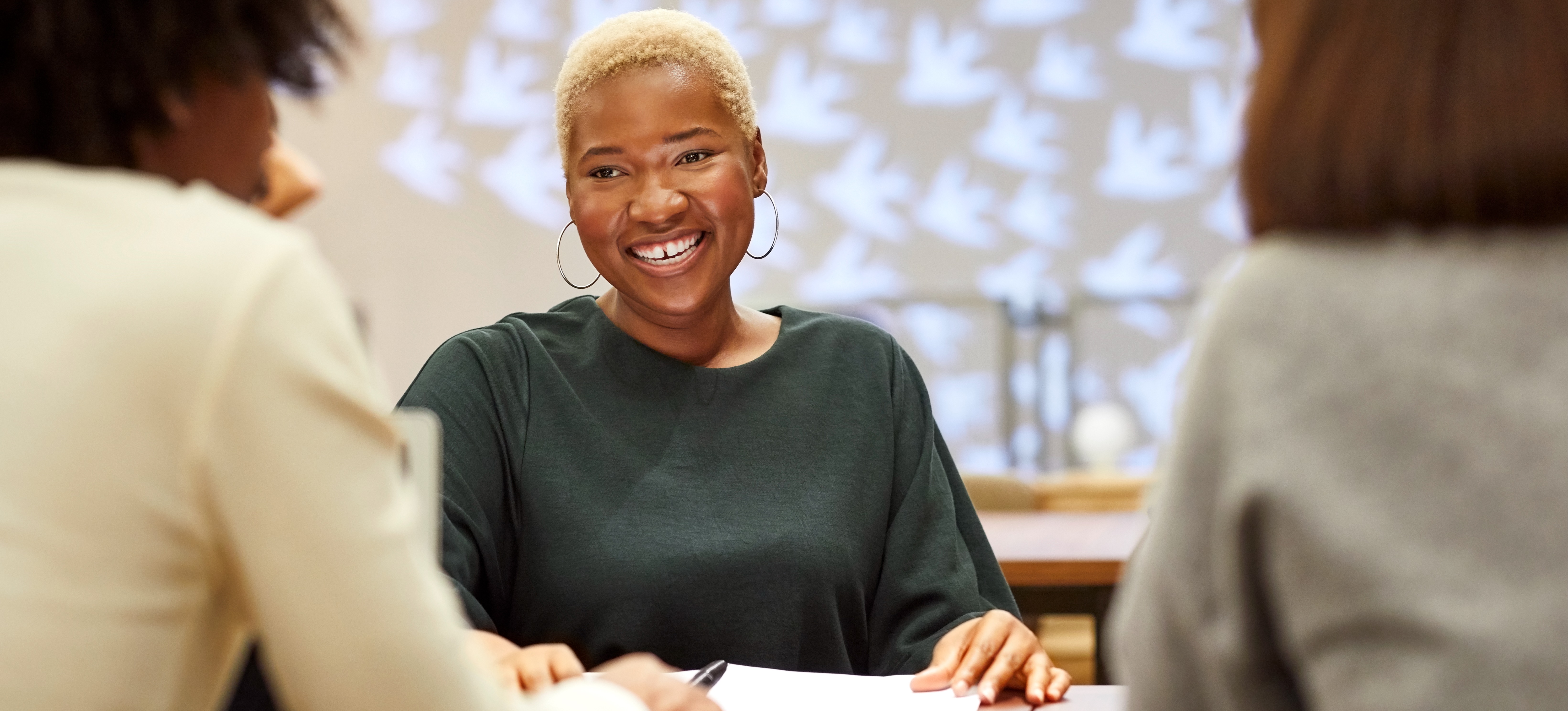[Featured Image] Two black people interview another black people who is smiling across the table with hoop earrings and green shirt.