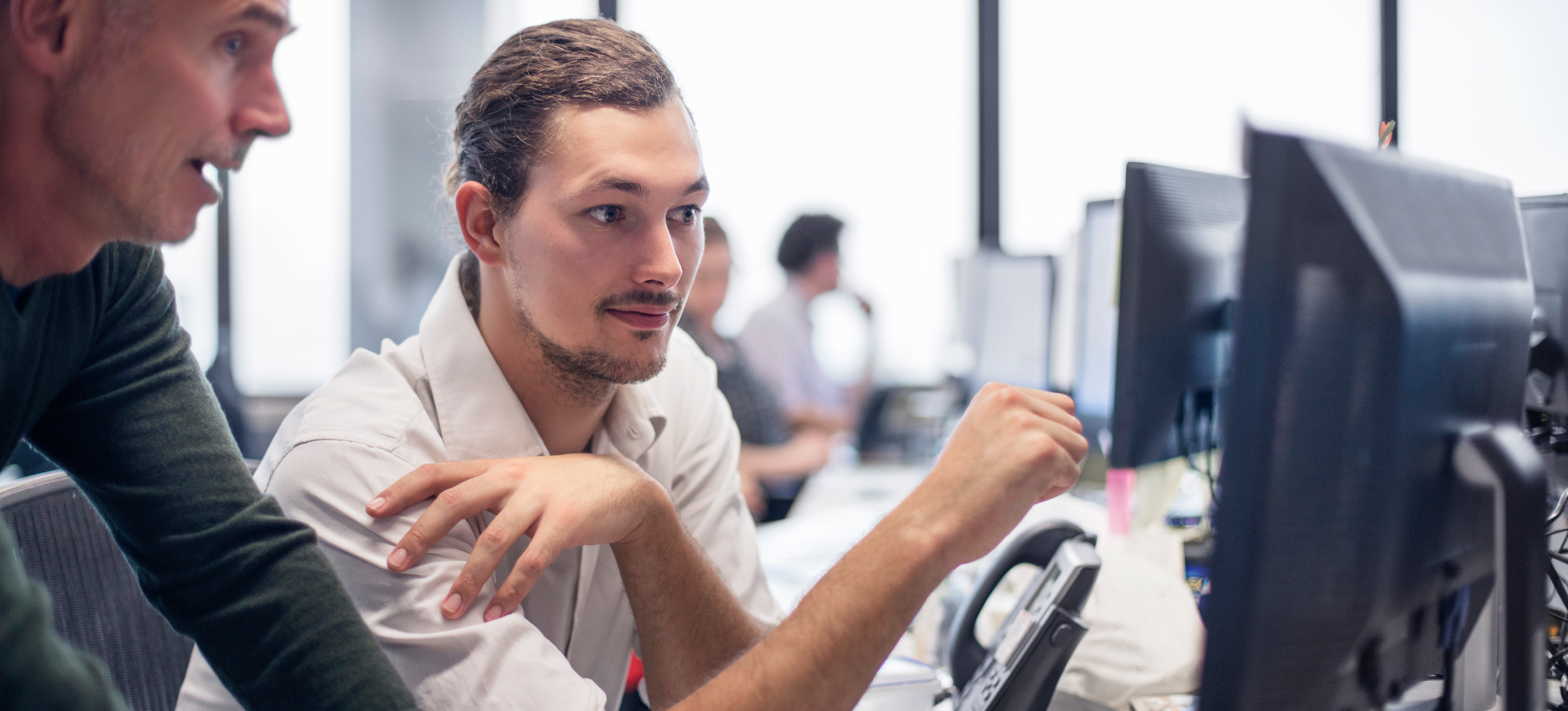 [Featured Image] Two programmers discuss what an epoch in machine learning is as they look at a computer screen in a spacious office that has several workstations and colleagues in it.
