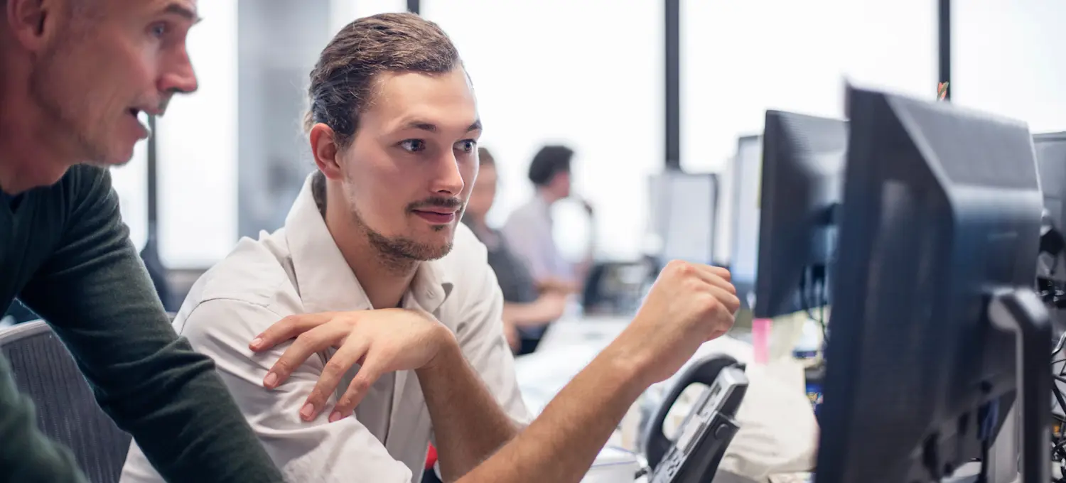 [Featured Image] Two programmers discuss what an epoch in machine learning is as they look at a computer screen in a spacious office that has several workstations and colleagues in it.
