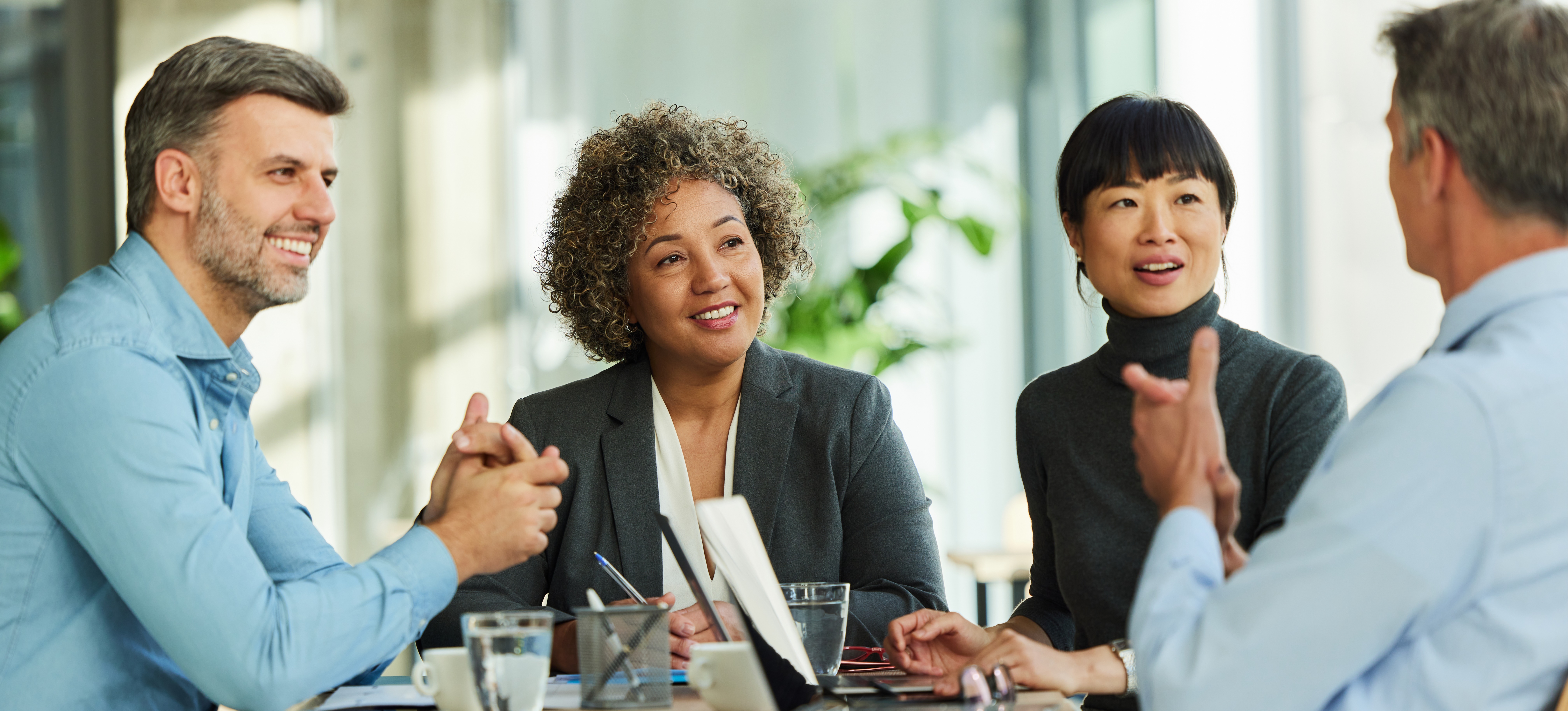 [Featured Image] Four colleagues sit at a table and discuss management interview questions they will ask their potential employees.
