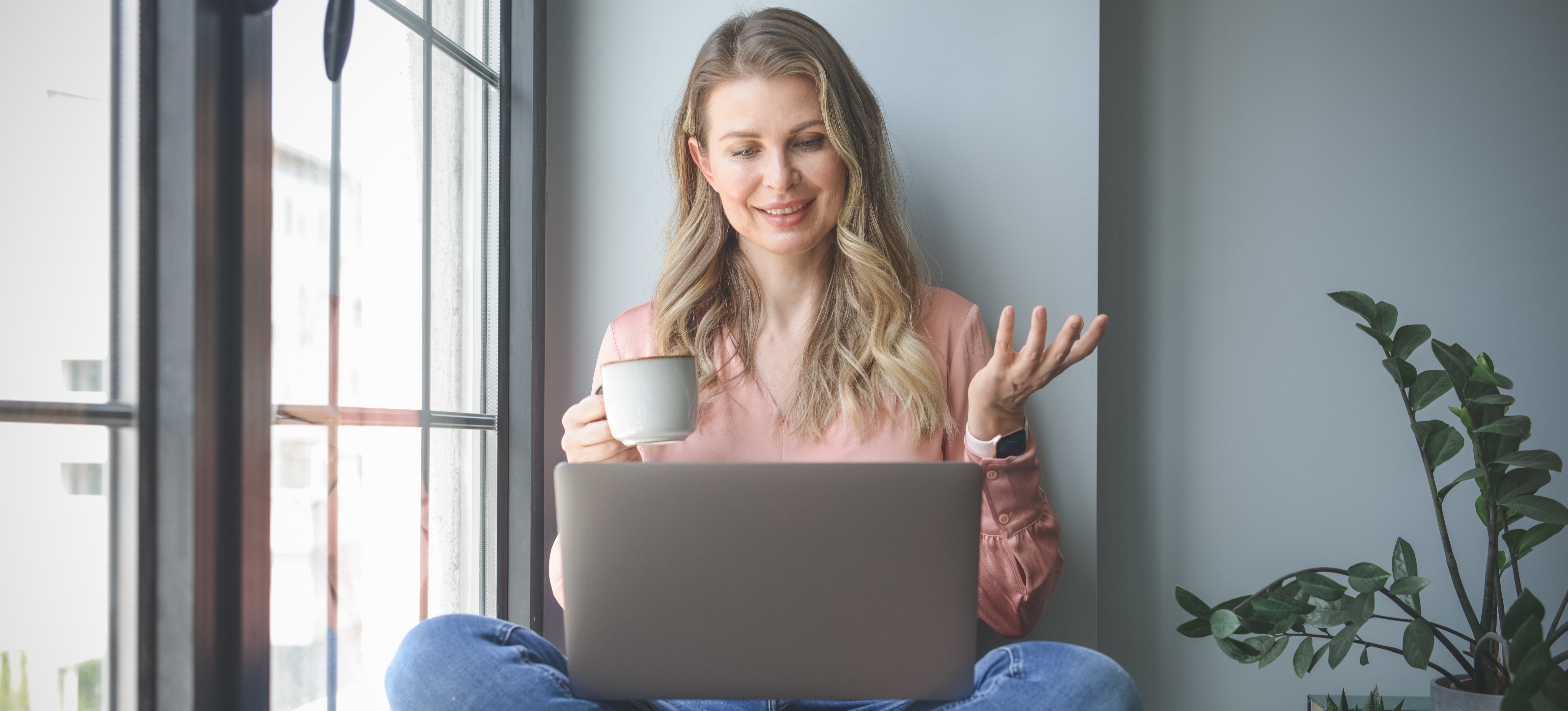 [Featured Image] A woman sits at home with a cup of coffee and her laptop and researches universities with low entry requirements. 
