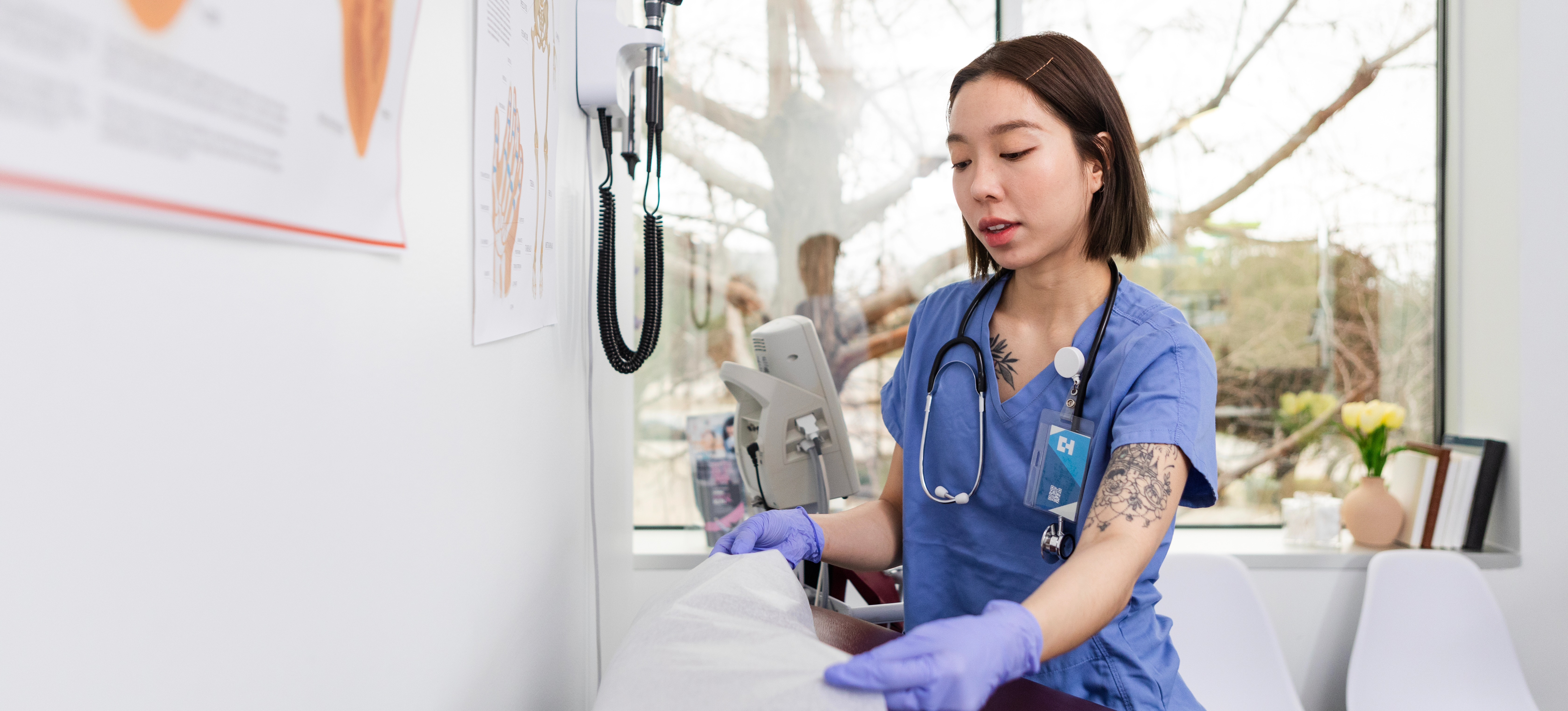 [Featured Image] A medical assistant wearing blue gloves and a stethoscope places paper over an exam table in a doctor's office.

