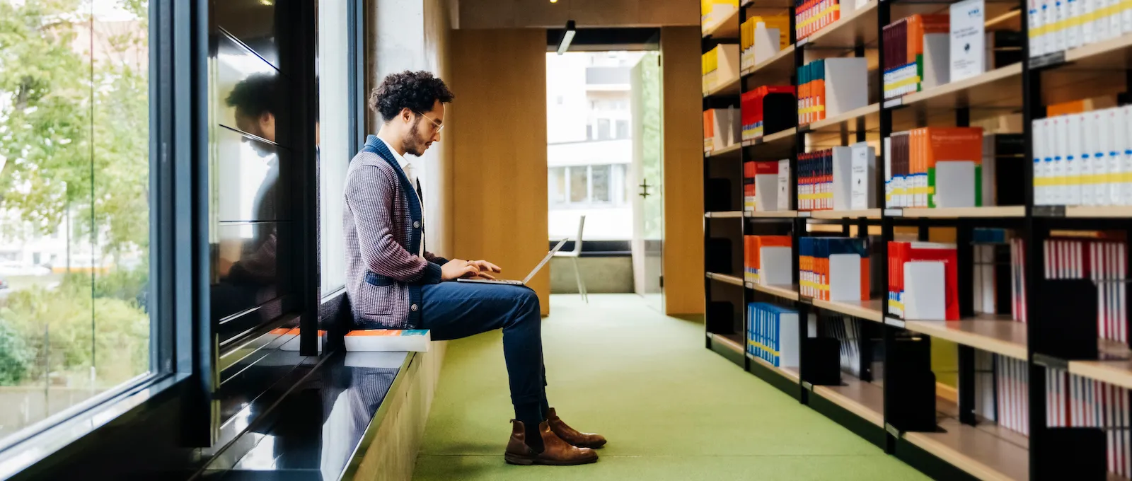 [Featured Image] View of a student sitting on the windowsill in a library, looking down at a laptop on their lap, across from shelves of reference books.