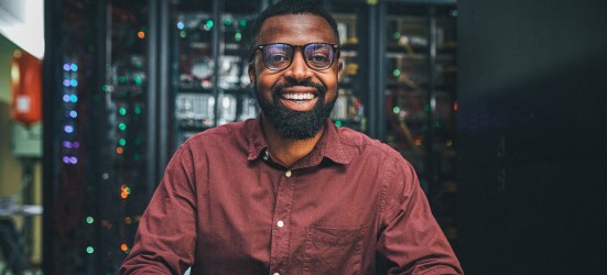 [Featured image] A Python developer in a maroon shirt stands in front of computer servers and smiles at the camera.
