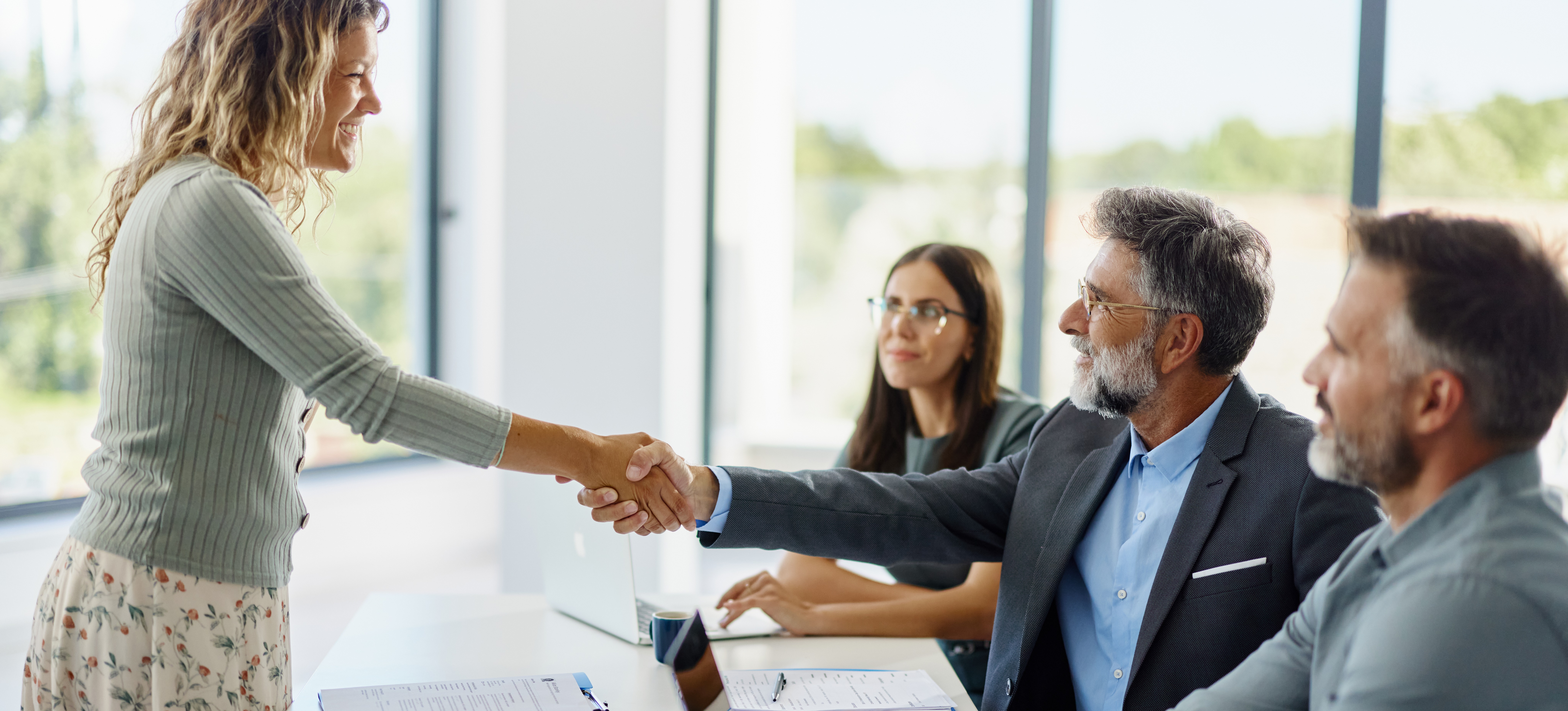 [Featured image] A smiling worker dressed in a skirt and sweater stands as they shake the hand of their interviewer after getting a job with no experience.
