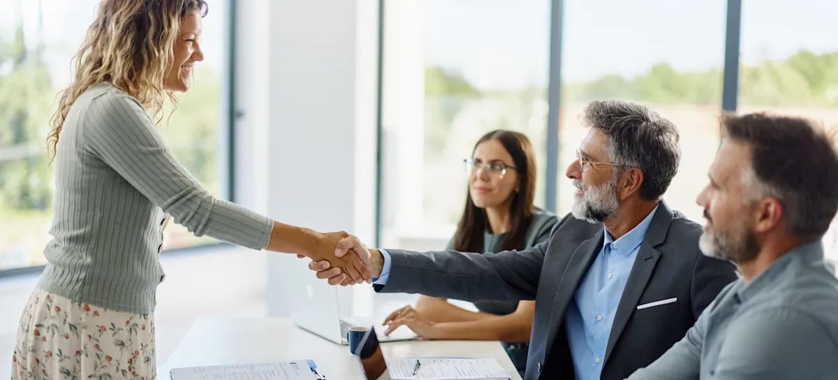 [Featured image] A smiling worker dressed in a skirt and sweater stands as they shake the hand of their interviewer after getting a job with no experience.
