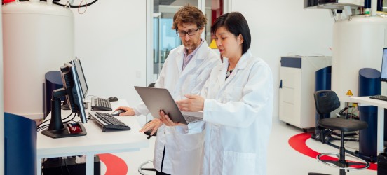 [Featured image] Two pharmacists in lab coats talk to each other while looking at a computer monitor.
