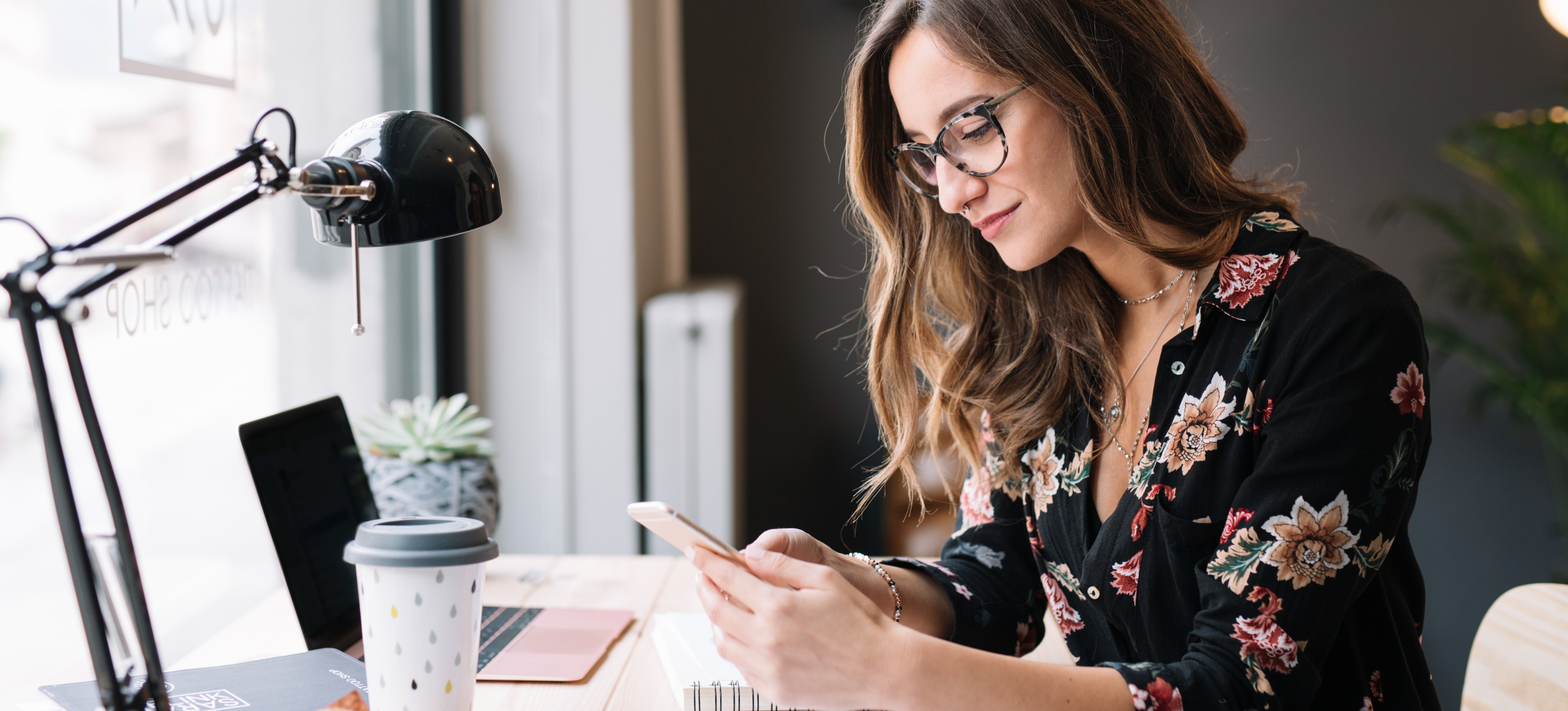 [Featured image] A person in a floral blouse sets up a dropshipping business on Shopify on their phone while sitting at a desk by a window.
