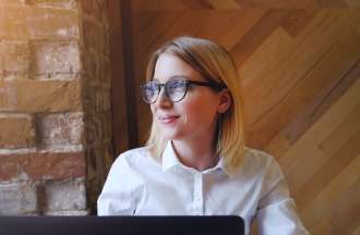 [Featured image]:  A female,  with blonde hair, wearing a white shirt and glasses, is sitting in front of her desktop working on an assignment as she works towards her business administration degree.