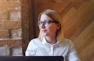 [Featured image]:  A female,  with blonde hair, wearing a white shirt and glasses, is sitting in front of her desktop working on an assignment as she works towards her business administration degree.