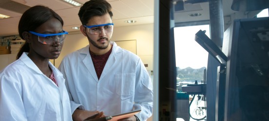 [Featured Image]: A health worker, with black hair, wearing a white uniform and blue goggles, and a person with black hair, wearing a white uniform and blue goggles. They are working in a lab, holding, writing and analyzing an experiment. 