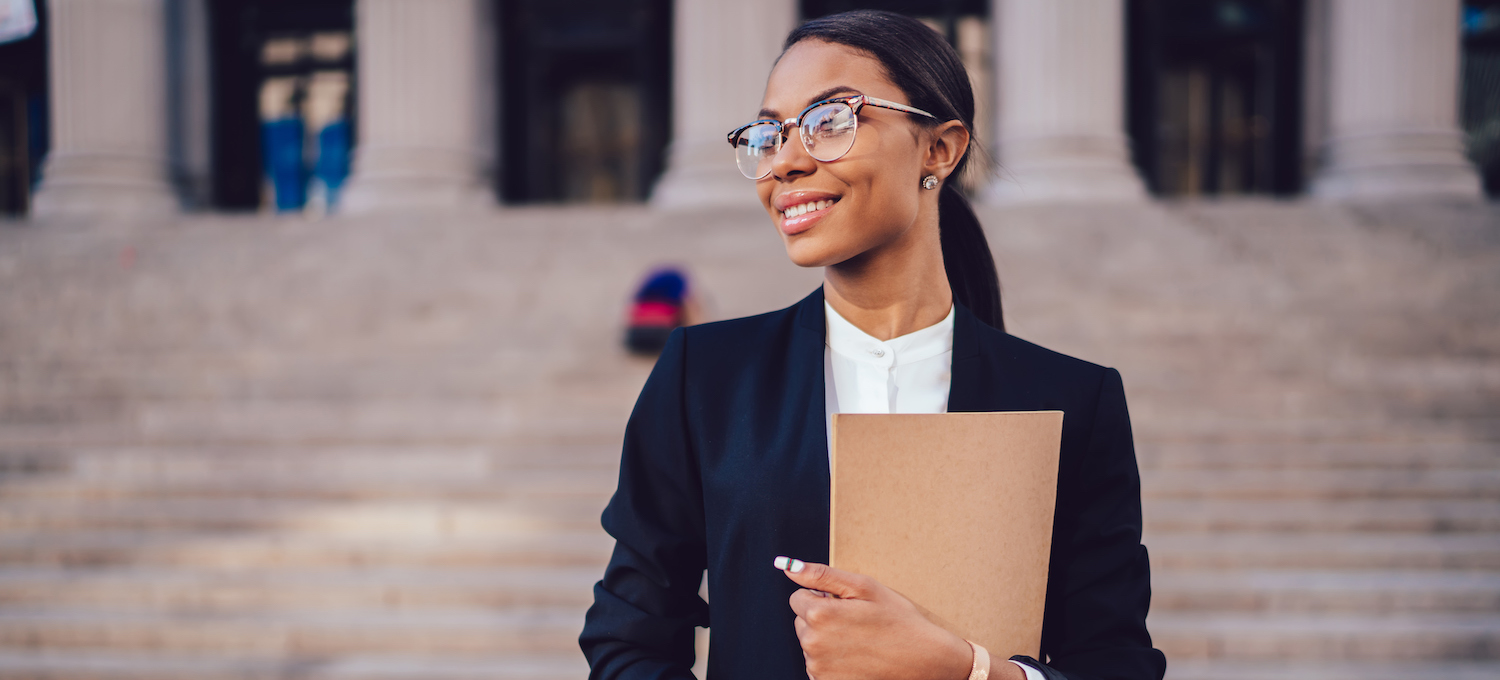 [Featured image] A JD degree candidate wearing glasses and a suit jacket stands in front of a building with pillars, holding a notebook.