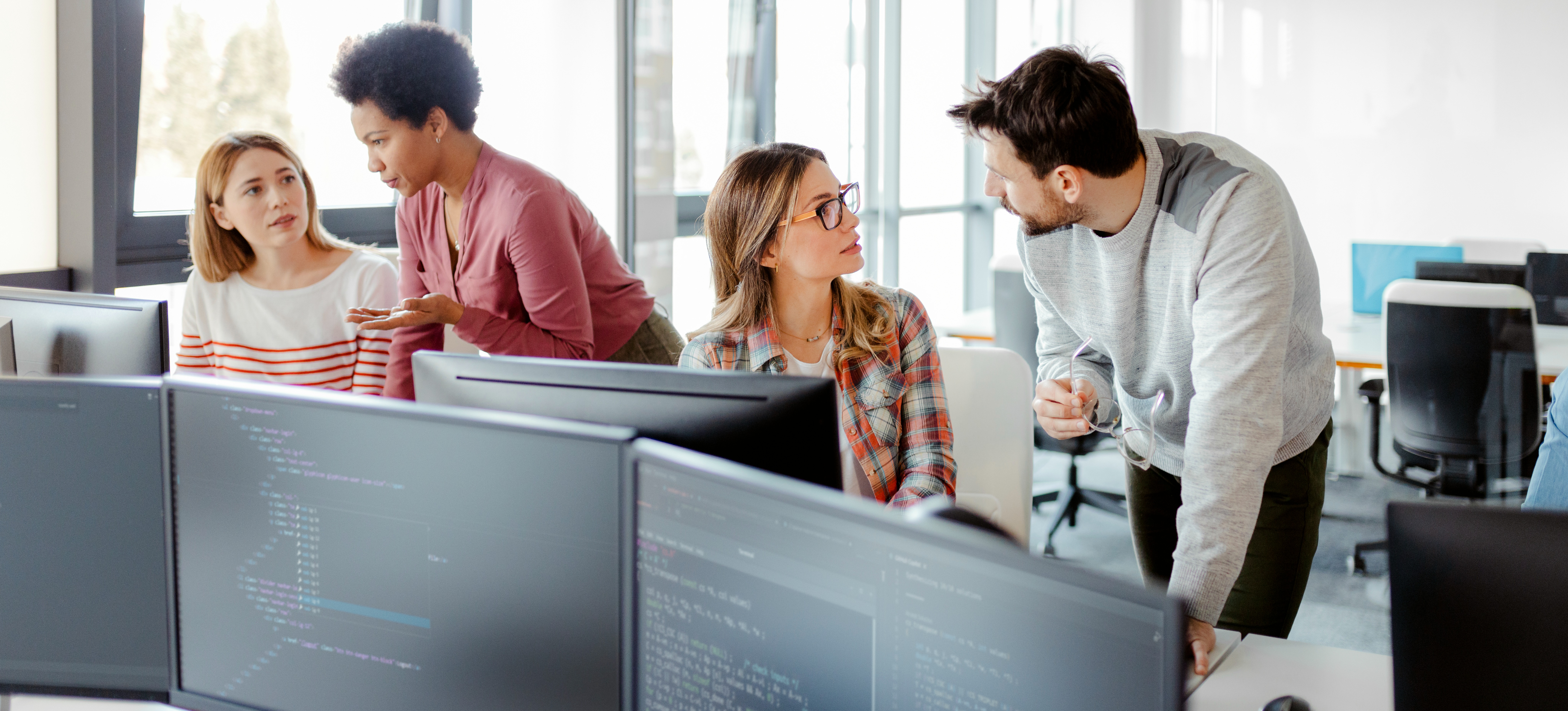 [Featured Image] Four professionals in an office sit and stand in front of several computer monitors and discuss DeepMind vs. OpenAI.
