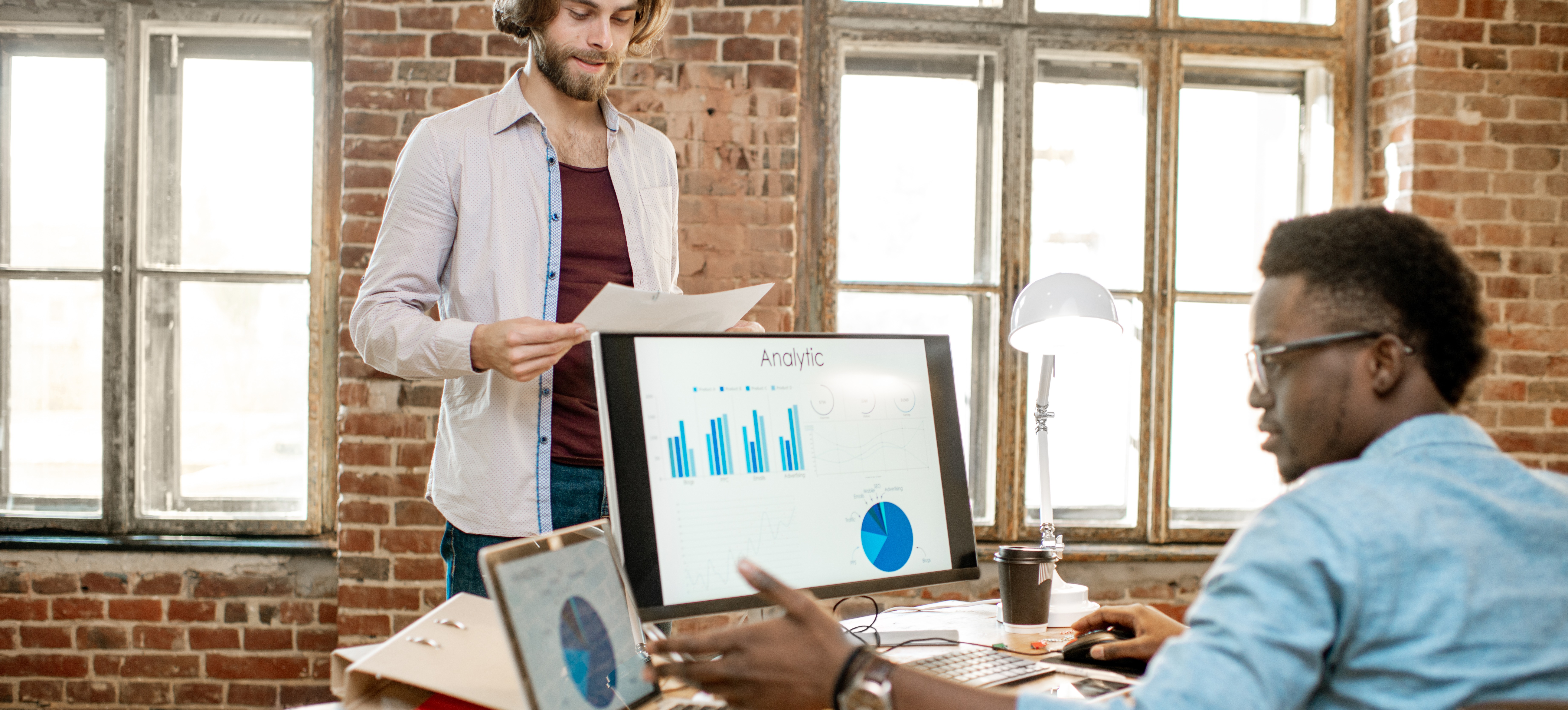 [Featured Image] Two market research analysts collaborate in a workspace, using market research methods.
