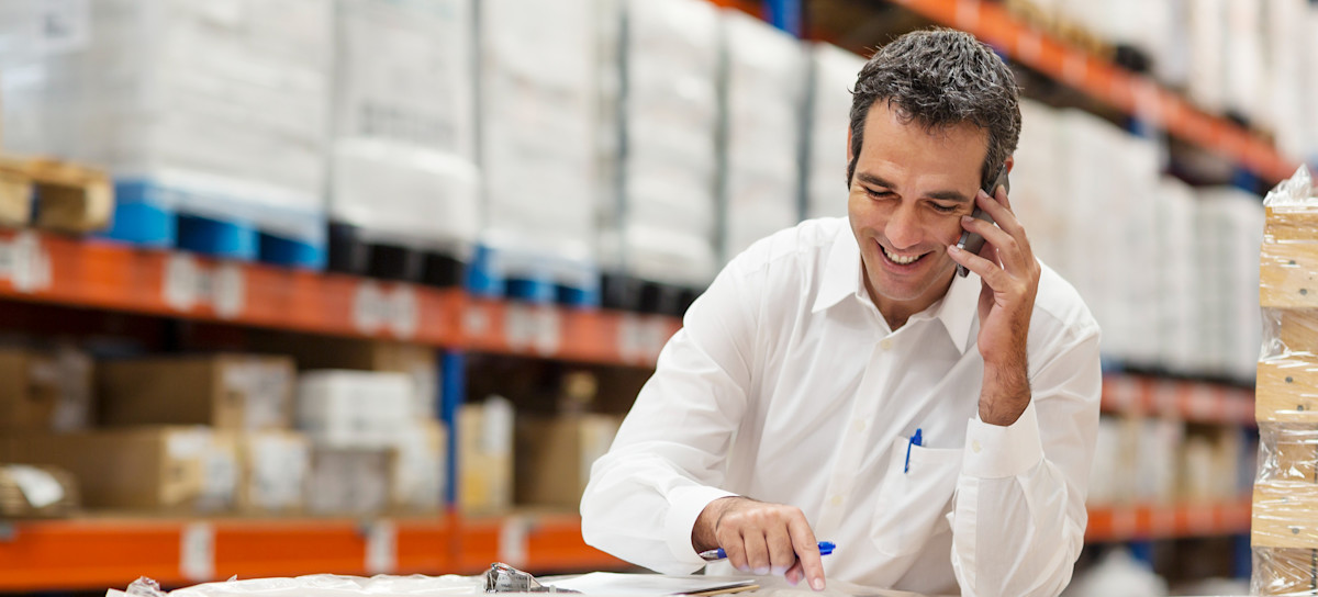 [Featured Image] A smiling professional uses product manager tools on their laptop as they stand at a table in a warehouse full of products.

