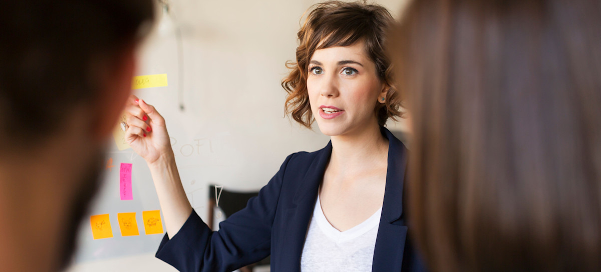 [Featured Image] A product marketing manager in a blazer is pointing to post-it notes while two coworkers are watching. 