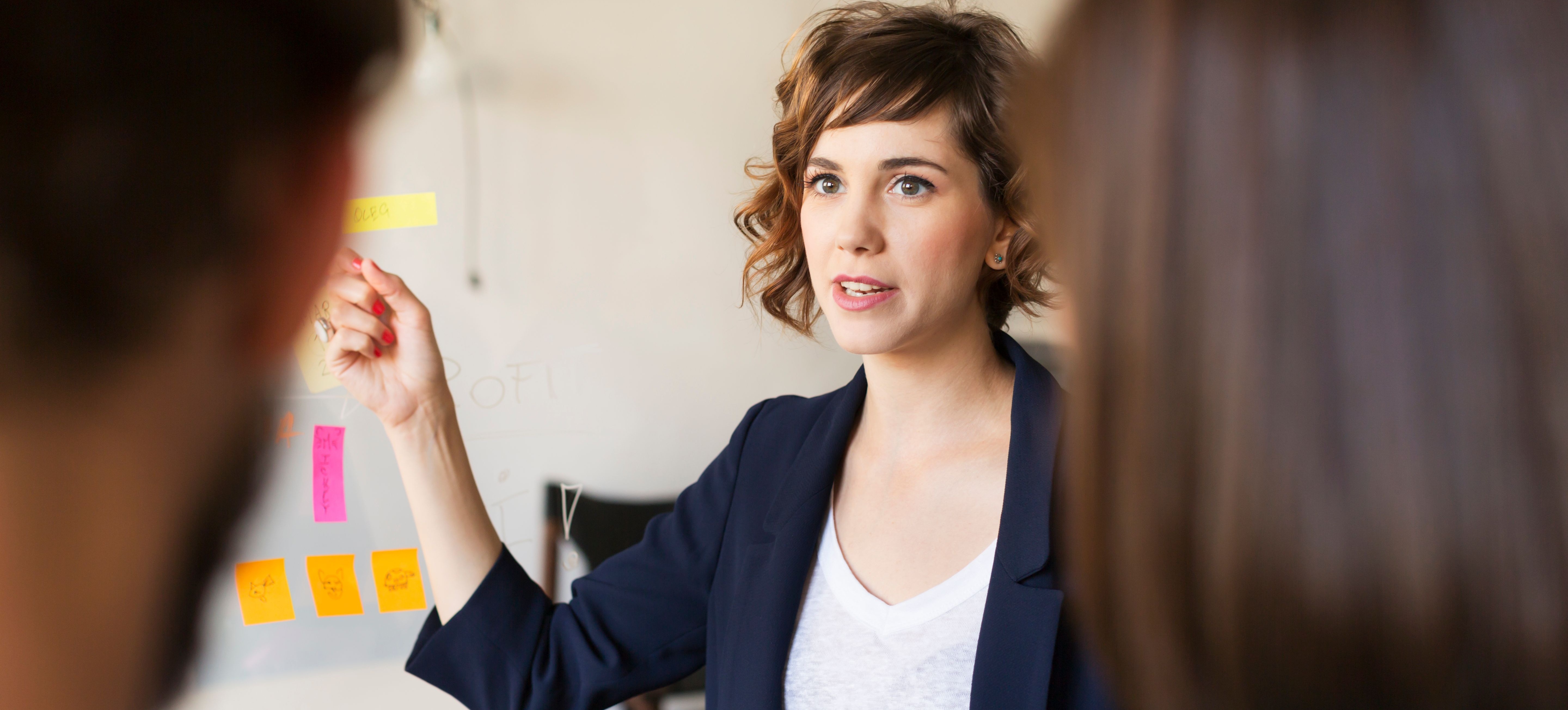 [Featured Image] A product marketing manager in a blazer is pointing to post-it notes while two coworkers are watching. 