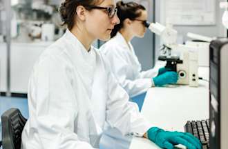 [Featured image] Two lab technicians in white coats and gloves work in a medical lab. One is typing on a computer and the other is looking at a specimen under a microscope.