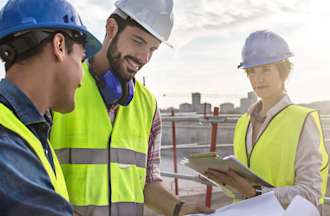 [Featured image] A construction manager reviews plans with two construction workers. They're all wearing hard hats and yellow work vests.