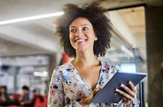 [Featured image] A product designer wears a floral blouse and stands in an open office holding a tablet. 