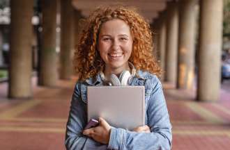[Featured image] A young white woman with curly orange hair stands outside in a portico holding a notebook and smiling. 