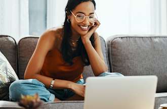 [Featured image] A smiling woman working from home sits on her grey sofa with her laptop on the coffee table in front of her.