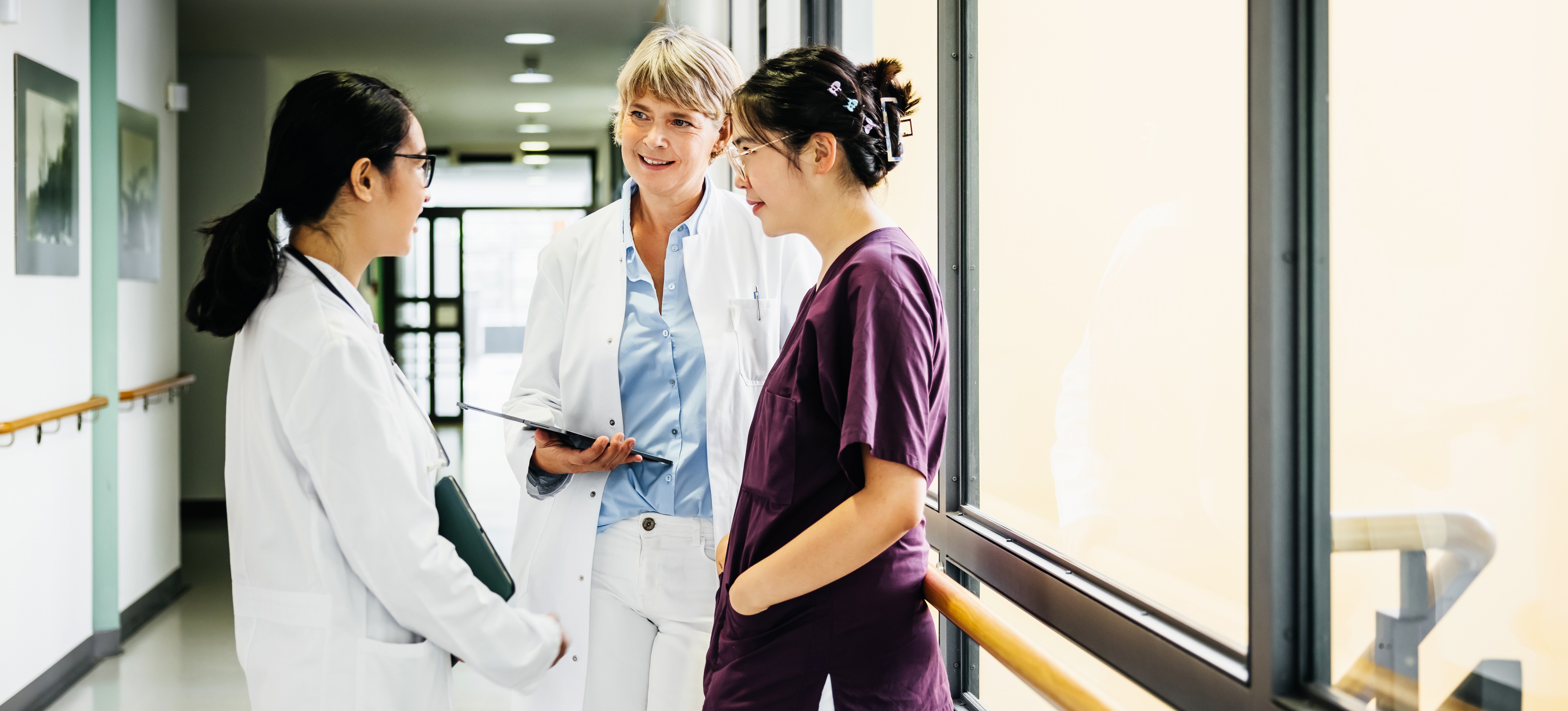 [Featured Image] Three medical professionals are talking about people skills in a wide hospital corridor with a bank of windows providing ample natural light to the right of them. 
