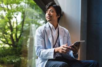 [Featured Image] A professional sits on a large window sill and gazes at green trees while holding a tablet.