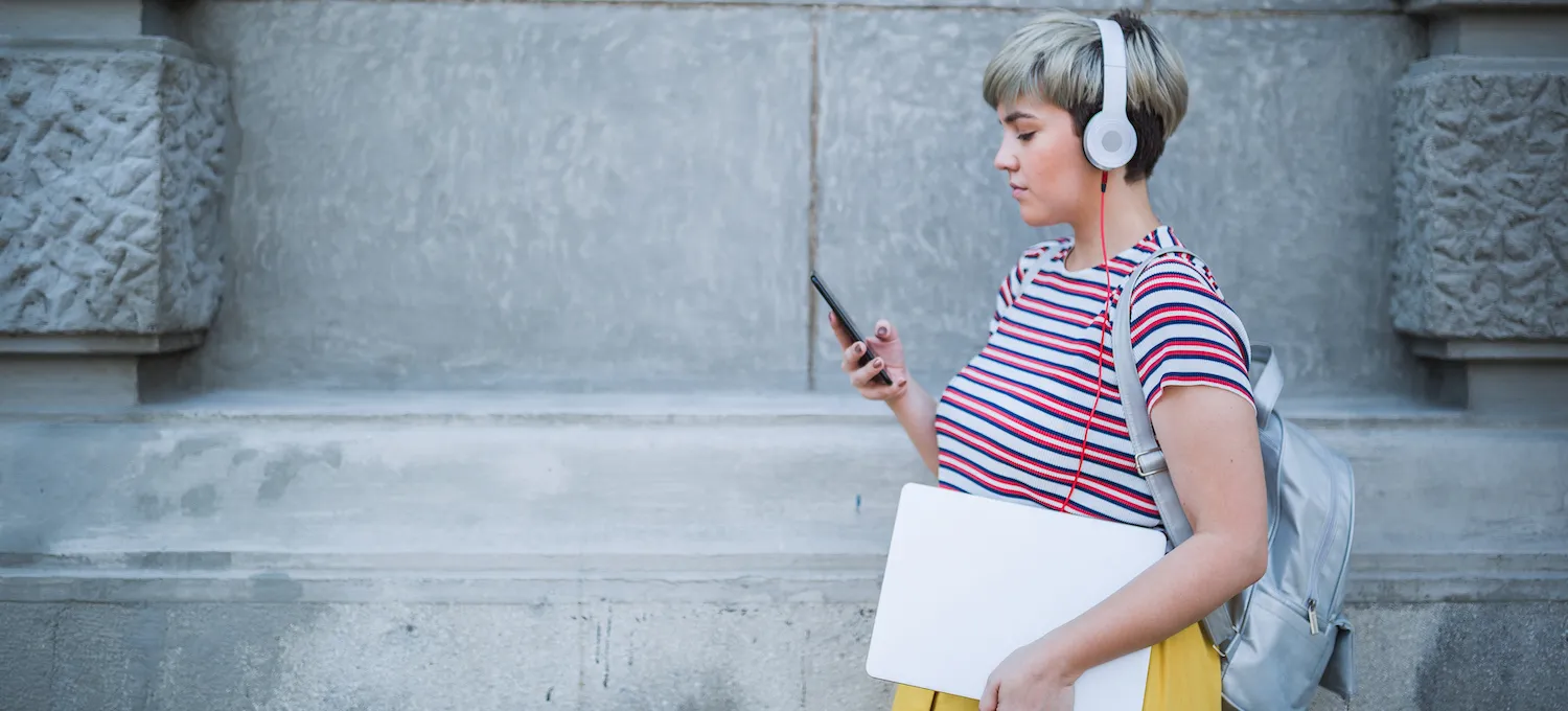 [Featured image] A young person wearing a striped shirt and listening to over-the-ear headphones walks while using their cell phone. 