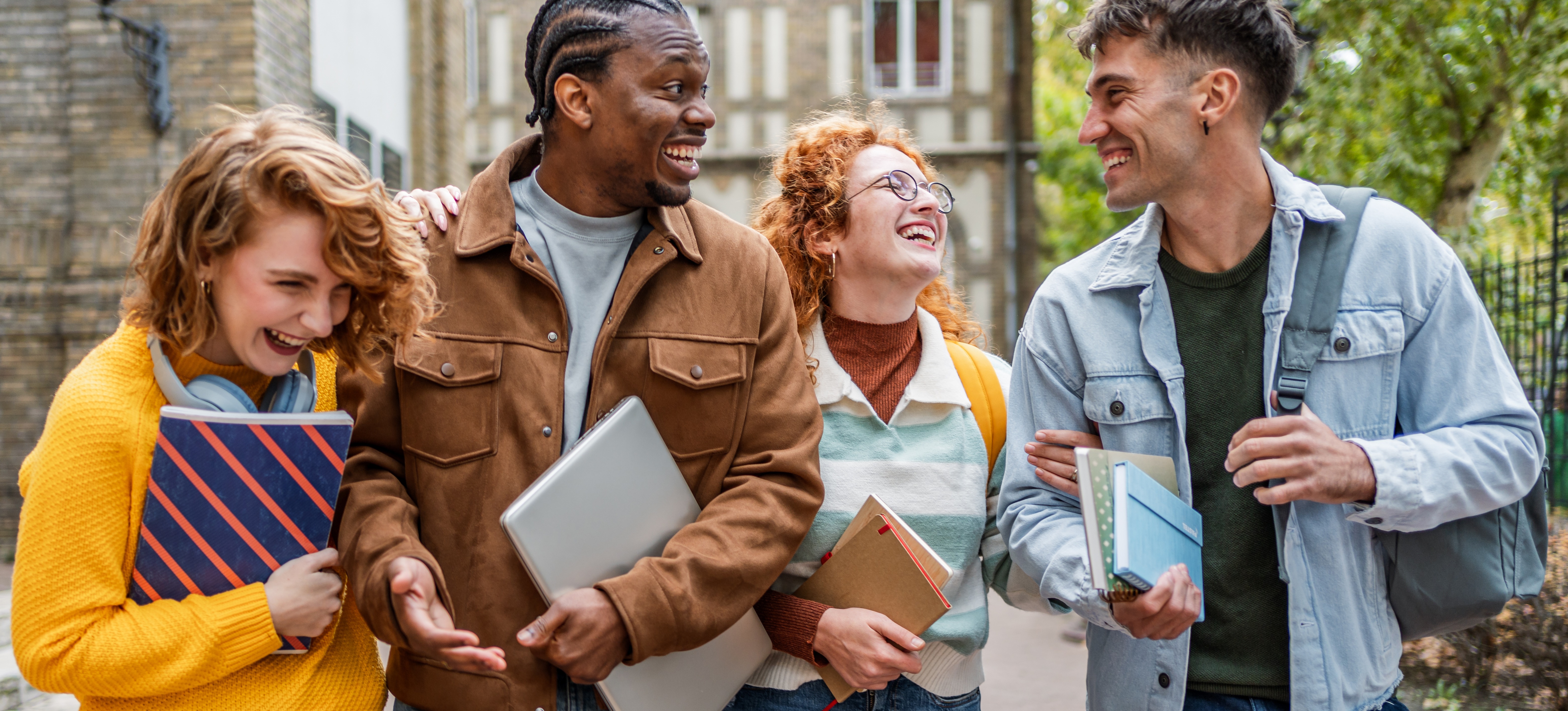 [Featured Image] Four students holding notebooks while smiling walk outside of a university campus.