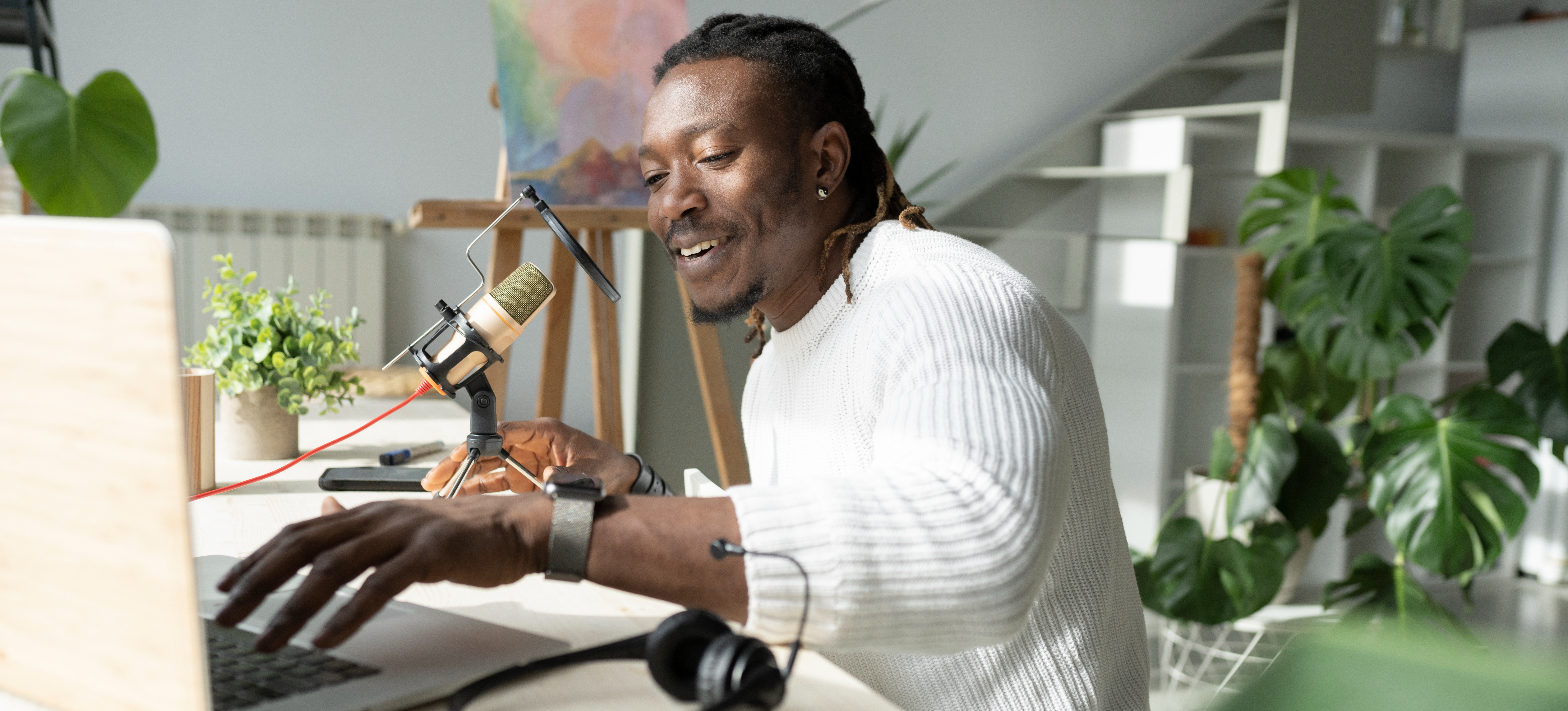 [Featured Image] A young professional uses a laptop and microphone for their side hustle after choosing to start a podcast from home in their spare time. 
