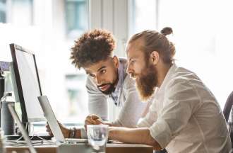 [Featured Image] Two AI professionals in an office setting looking at computer screens discuss artificial general intelligence vs. AI. 